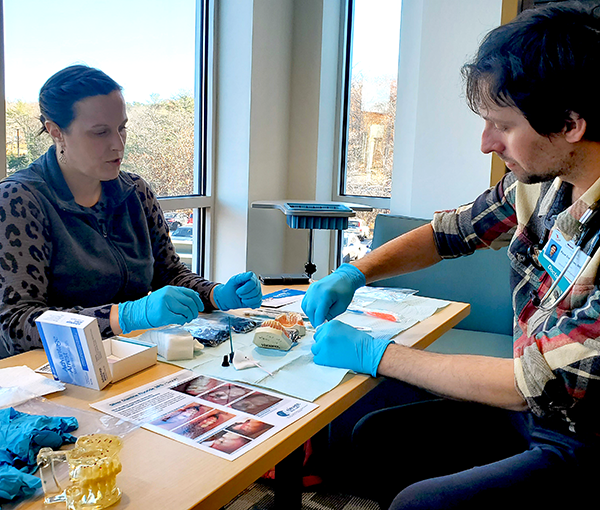 Woman on side of desk offering training to man on the other wearing gloves and utilizing dentistry tools.