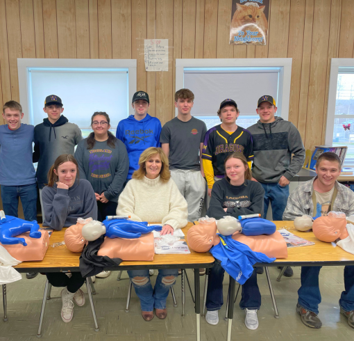 A group of high school students posing for the camera with CPR dummies on a table