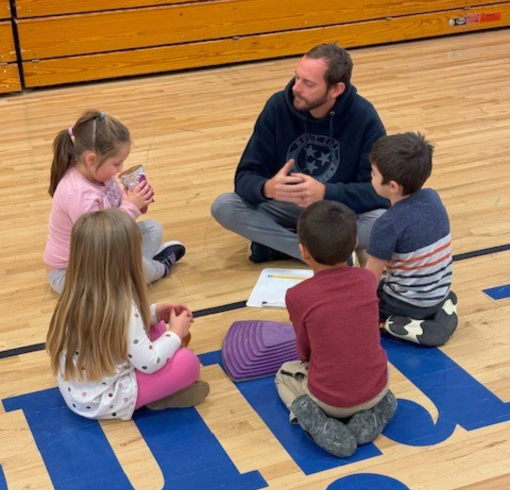 A young male teacher sitting on the floor of a gym in a circle with chidlren.