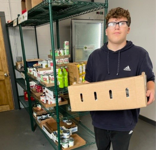 A young man holding a box in front of shelves of food in a school.