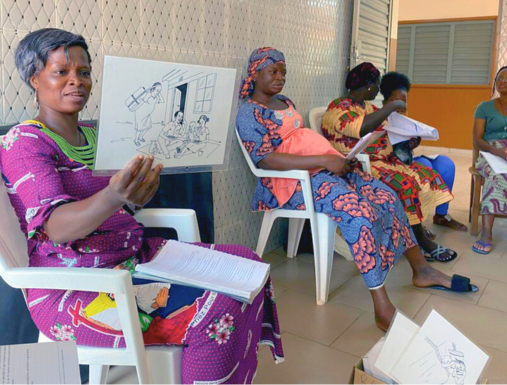 A woman in a dress holds up a sign while talking to a group of pregnant women in Benin.