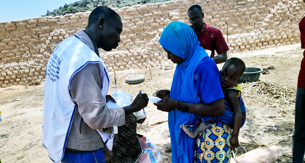 A man wearing a white official vest talks with a woman in a blue headscarf carrying a child on her back outside.