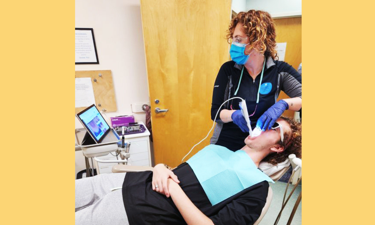 Woman wearing a blue mask uses a dental camera to examine a boy's teeth while sitting in a dental chair in an office