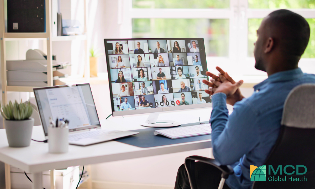 A man sitting in a chair looking at a screen with numerous people on it who are all on a meeting.