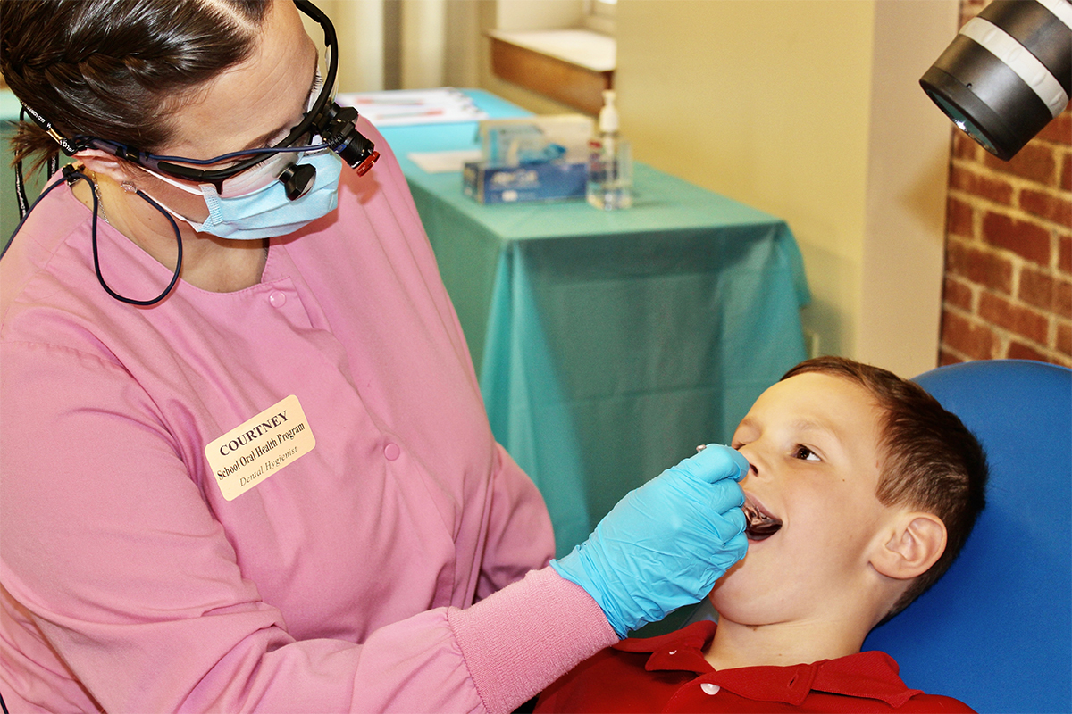 Courtney Vannah in pink scrubs with mask and glasses examines a young boys teeth.