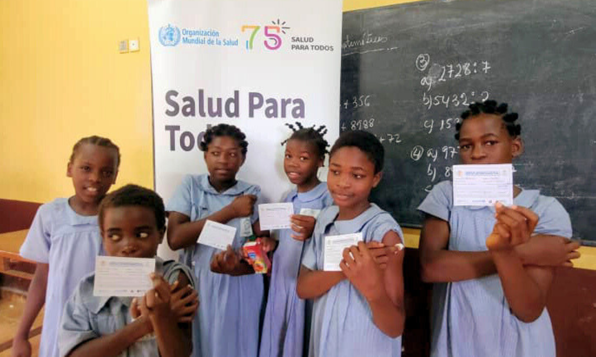 Group of young girls in blue dresses in front of school chalkboard holding up new vaccine cards after getting the HPV vaccine in Equatorial Guinea.