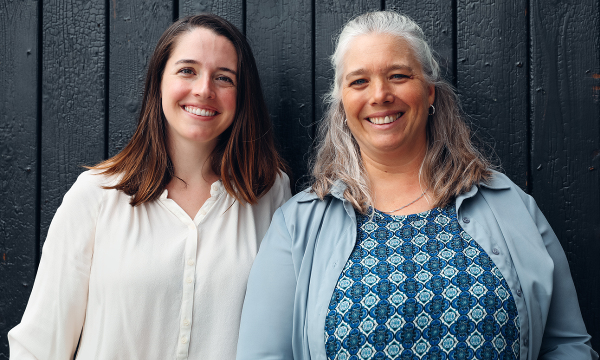 Photo of two women smiling at camera in front of blue wood background.