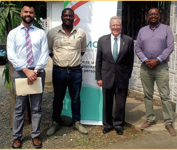 Four men standing for the camera in front of an MCD banner outside in Equatorial Guinea who are part of the supply chain management project.