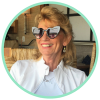 Woman with blonde hair smiling at camera while wearing heart-shaped sunglasses outside.