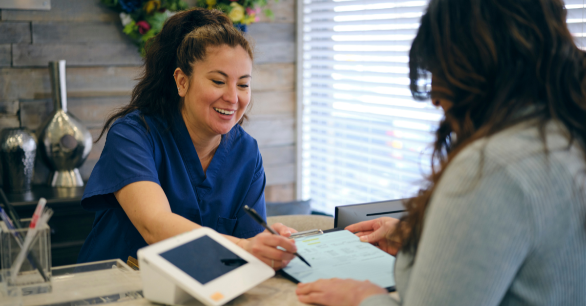 Smiling lady in scrubs helping a patient check out at a clinic in a bright setting.