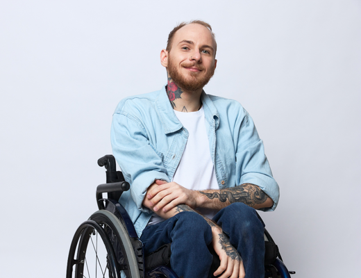A smiling man who uses a wheelchair, wearing a denim jacket.