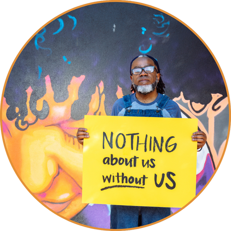 A Deaf Black man wearing glasses looks neutrally at the camera while holding a hand lettered sign declaring, “NOTHING about us without US.